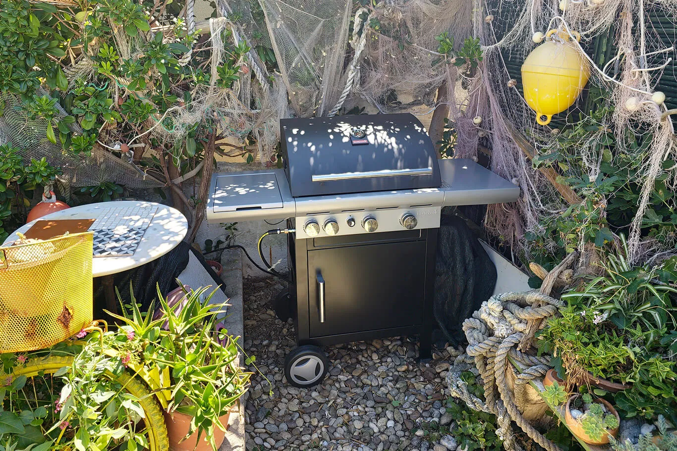 A four-burner gas BBQ surrounded by fishing nets, buoys and rocks